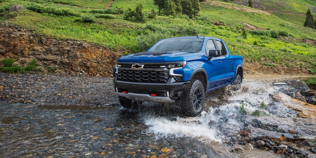 Image of a new blue Chevrolet Silverado driving through a creek at the base of a mountain on a sunny day 